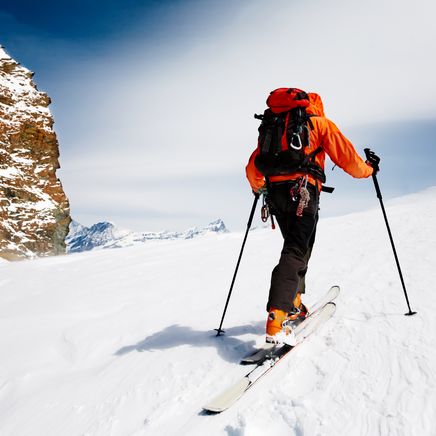 Un homme de dos en ski rando, et un blouson orange et son sac à dos, accompagné de ses harnais est en train de monter une pente vertigineuse.