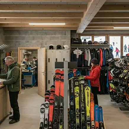 Intérieur d'un magasin de location de ski : clients au comptoir et devant des racks de skis, bottes, vêtements, avec vue sur des pistes enneigées.