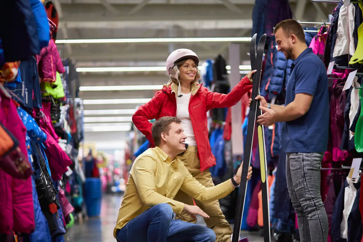 Un couple souriant essaie des skis avec un vendeur dans un magasin de sport, entouré de vêtements d'hiver.