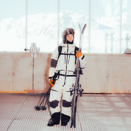 Skieur en tenue blanche, casque et gants orange, tenant skis et bâtons, dans un bâtiment vitré avec vue sur montagnes enneigées.