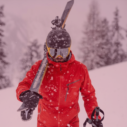 Skieur en veste rouge, casque et masque, portant ses skis sur l'épaule et des bâtons, sous une chute de neige en montagne.