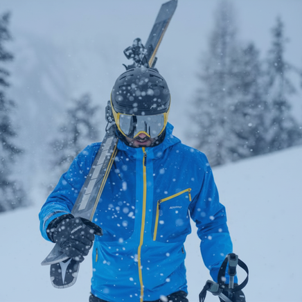 Un skieur équipé d'un casque, d'un masque et d'une veste bleue, portant des skis sur l'épaule et des bâtons, marche sous la neige dans un paysage hivernal.