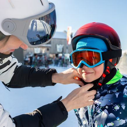 Une personne ajuste le casque d'un enfant souriant et équipé pour le ski, sur une piste enneigée et ensoleillée.